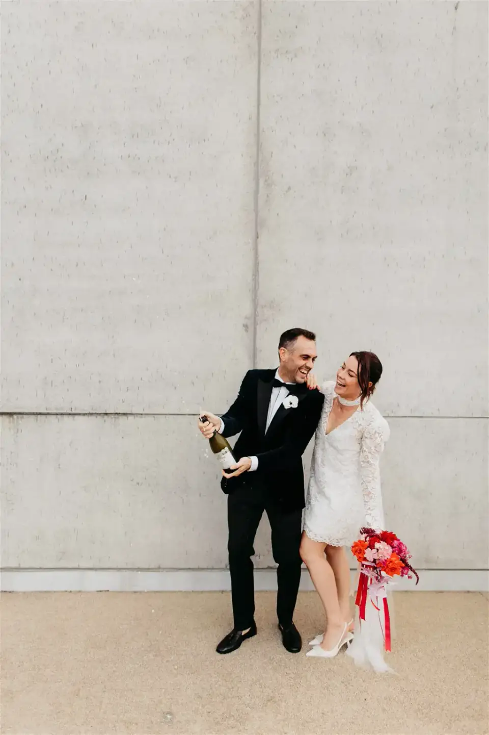 Christian and Amy sheltering under an umbrella while sharing an intimate moment against The Calile's textured exterior wall